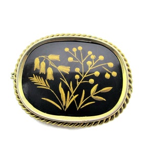 Puede incluir: Un broche ovalado de color dorado con fondo de esmalte negro y un dise&ntilde;o floral dorado.