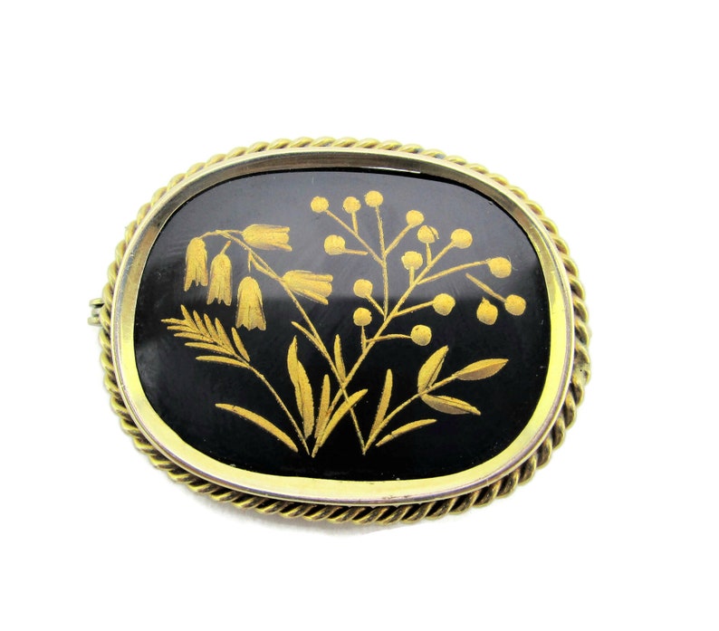 Puede incluir: Un broche ovalado de color dorado con fondo de esmalte negro y un dise&ntilde;o floral dorado.