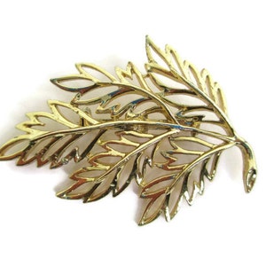 May include: A gold-toned metal brooch shaped like a leafy branch. The brooch is made of delicate, intricate cutouts that resemble leaves.