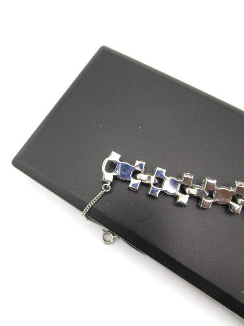 Puede incluir: Un brazalete plateado con un dise&ntilde;o de cadena y brillantes transparentes. El brazalete est&aacute; sobre una superficie negra.