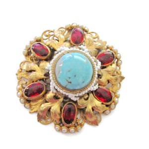 May include: A gold-toned brooch with a turquoise cabochon surrounded by smaller red glass stones and white pearls.