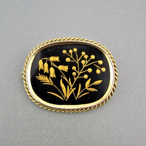 Puede incluir: Un broche ovalado en tono dorado con fondo de esmalte negro y un dise&ntilde;o floral detallado en oro. Las flores son estilizadas y tienen un aspecto delicado e intrincado.