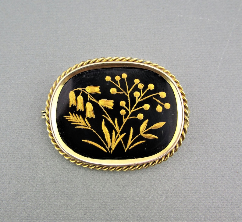 Puede incluir: Un broche ovalado en tono dorado con fondo de esmalte negro y un dise&ntilde;o floral detallado en oro. Las flores son estilizadas y tienen un aspecto delicado e intrincado.