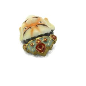 May include: A small ceramic figurine of a face with a colorful, ornate collar. The face is painted with orange, white, and black details. The collar is decorated with red, blue, and gold accents.