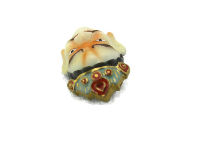 May include: A small ceramic figurine of a face with a colorful, ornate collar. The face is painted with orange, white, and black details. The collar is decorated with red, blue, and gold accents.