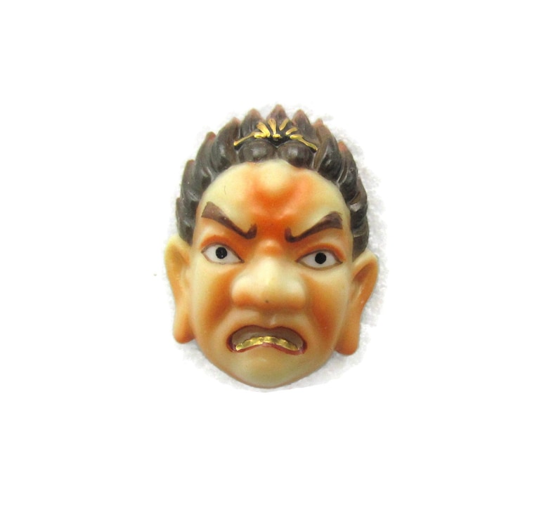 May include: A ceramic mask with a stylised human face. The mask has brown and black hair, a light orange face, and a gold painted mouth. The mask has a frowning expression.