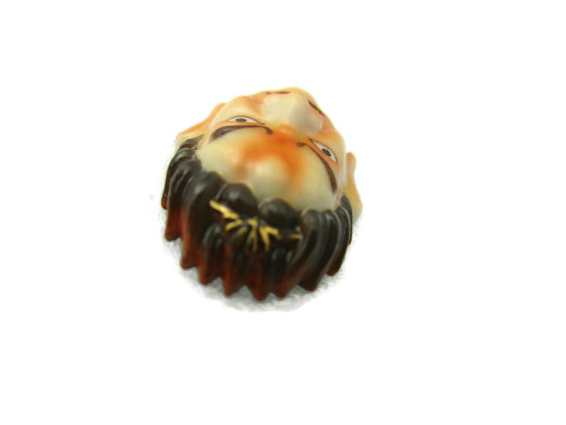 May include: A ceramic figurine of a person's face with a brown beard and moustache. The face is painted with orange, white and black details.