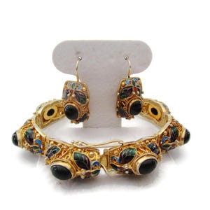 Könnte beinhalten: Ein goldenes Armband und passende Ohrringe mit schwarzen Edelsteinen und bunten floralen Emaille-Designs.