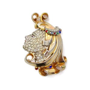 May include: A gold tone brooch with a stylized face design, set with clear rhinestones and colorful enamel accents.