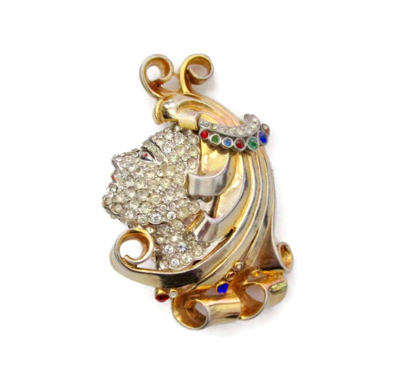 May include: A gold tone brooch with a stylized face design, set with clear rhinestones and colorful enamel accents.