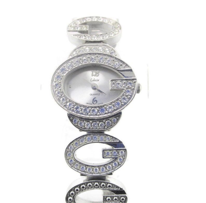 May include: Silver-toned wristwatch with a white face and crystal accents. The watch face features the text "Colezer Quartz" and the band is made up of interlocking "G" letters.