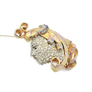 May include: A gold-tone brooch with a faceted crystal design. The brooch features a stylized face with a ribbon design and colorful accents.