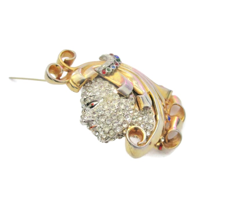 May include: A gold-tone brooch with a faceted crystal design. The brooch features a stylized face with a ribbon design and colorful accents.