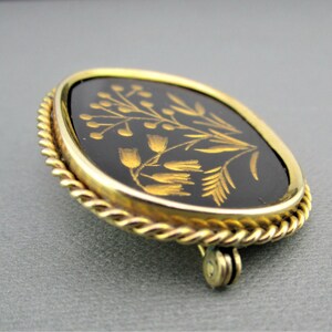 Puede incluir: Un broche dorado con un camafeo ovalado negro que presenta un dise&ntilde;o floral en oro. El camafeo est&aacute; rodeado por un borde de cuerda.