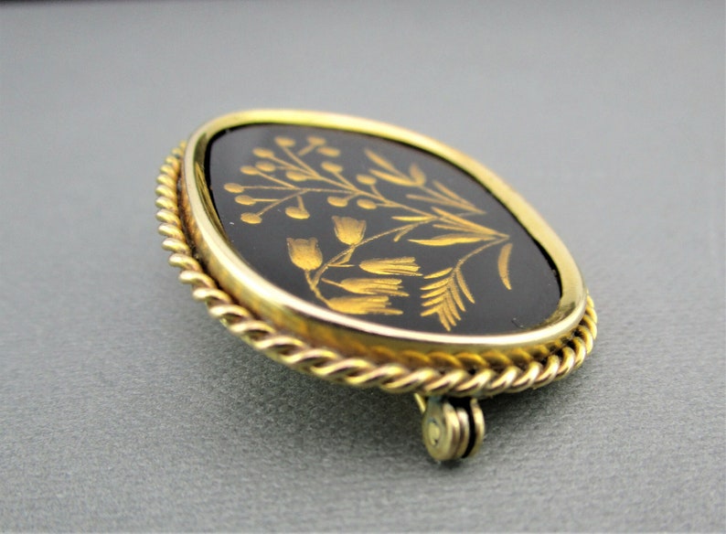 Puede incluir: Un broche dorado con un camafeo ovalado negro que presenta un dise&ntilde;o floral en oro. El camafeo est&aacute; rodeado por un borde de cuerda.