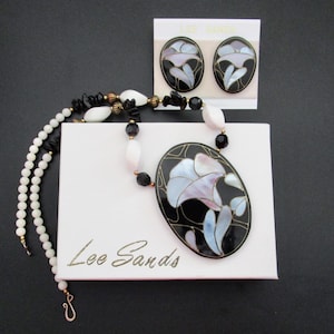 May include: A vintage jewelry set featuring a necklace and matching earrings. The necklace has a black oval pendant with a floral design in white, pink, and blue. The earrings and pendant are on a white box with the text "Lee Sands".