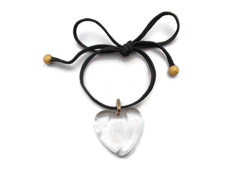 May include: A clear heart-shaped crystal pendant hangs from a black cord necklace with gold-toned beads at the ends.