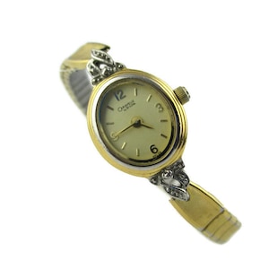 May include: A gold-tone wristwatch with a white face and black Roman numeral hour markers. The watch has a gold-tone band with a decorative design on the clasp. The watch face says "Caravelle by Bulova".