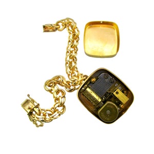May include: A gold chain bracelet with a square music box charm. The music box is open, revealing the inner workings of the mechanism.