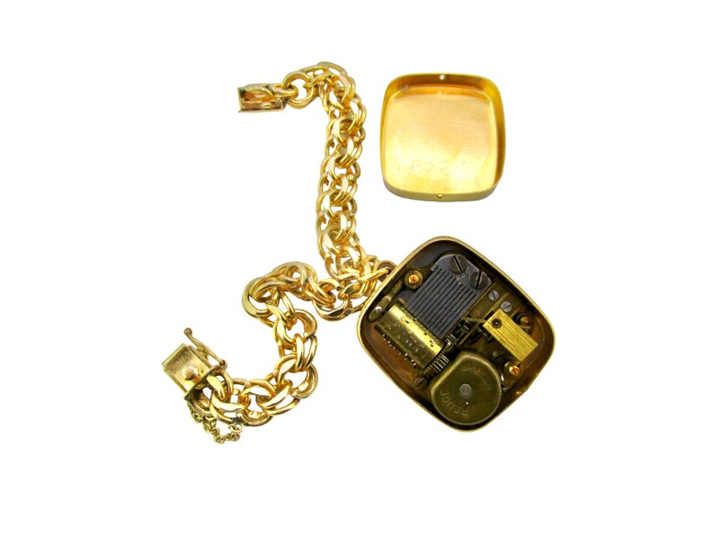 May include: A gold chain bracelet with a square music box charm. The music box is open, revealing the inner workings of the mechanism.