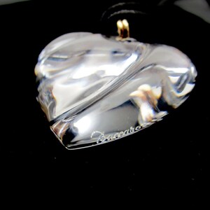 May include: A clear crystal heart pendant necklace with a gold chain and the word "Baccarat" engraved on the heart.
