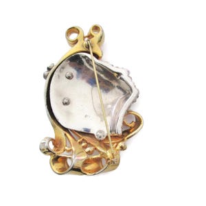 May include: A gold and silver tone brooch with a faceted stone design. The brooch has a pin and catch closure on the back.