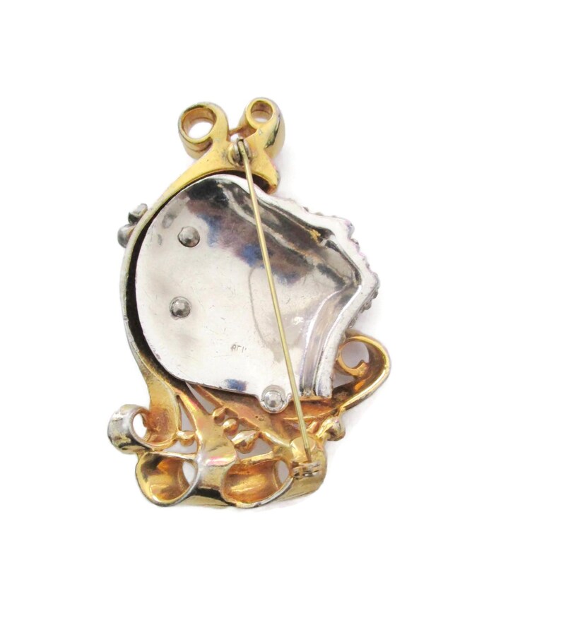 May include: A gold and silver tone brooch with a faceted stone design. The brooch has a pin and catch closure on the back.