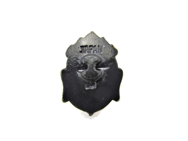 May include: Black metal pin with a detailed design. The back of the pin has the word "JAPAN" printed on it.