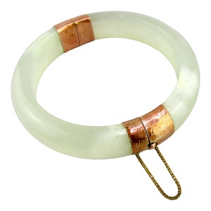 May include: A pale green jade bangle bracelet with copper-colored metal accents. The bracelet is round and has a clasp with a small gold chain. The jade has a translucent quality.