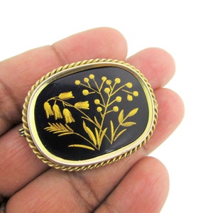 Puede incluir: Un broche dorado con fondo de esmalte negro y un dise&ntilde;o floral detallado en oro. Las flores son estilizadas y tienen un aspecto delicado e intrincado.
