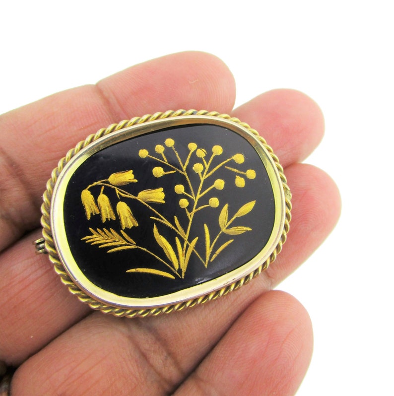Puede incluir: Un broche dorado con fondo de esmalte negro y un dise&ntilde;o floral detallado en oro. Las flores son estilizadas y tienen un aspecto delicado e intrincado.