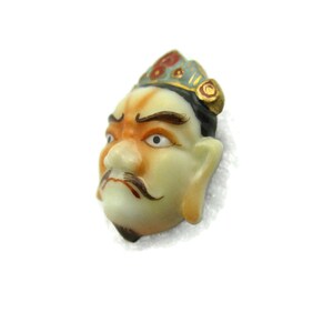 May include: A porcelain figurine of a man's face with a crown, painted in shades of green, red, and gold. The face has a serious expression with a mustache and beard.
