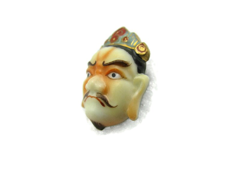 May include: A porcelain figurine of a man's face with a crown, painted in shades of green, red, and gold. The face has a serious expression with a mustache and beard.