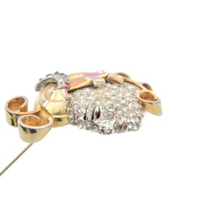May include: A gold-tone brooch with a large, clear rhinestone center and smaller rhinestones surrounding it. The brooch has a gold-tone pin and a decorative scroll design on the side.