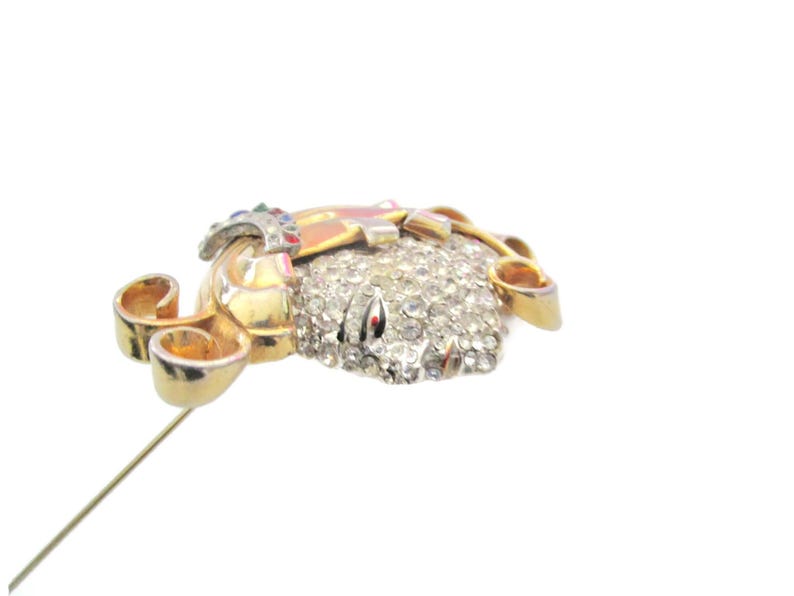 May include: A gold-tone brooch with a large, clear rhinestone center and smaller rhinestones surrounding it. The brooch has a gold-tone pin and a decorative scroll design on the side.
