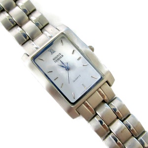 May include: A silver watch with a rectangular face and a silver metal band. The watch face is white with black Roman numeral hour markers and black hands. The watch face also has the words "Bijoux Terner" and "Quartz" printed on it.
