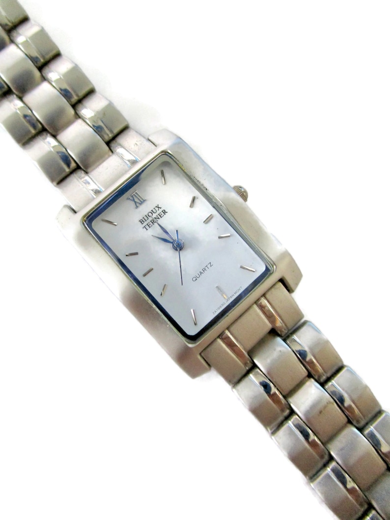 May include: A silver watch with a rectangular face and a silver metal band. The watch face is white with black Roman numeral hour markers and black hands. The watch face also has the words "Bijoux Terner" and "Quartz" printed on it.