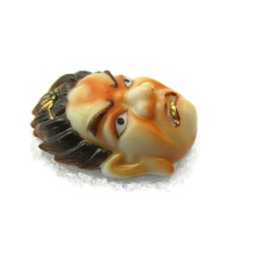 May include: A ceramic mask with a detailed face, painted in shades of orange and brown. The mask has black hair and gold accents around the eyes and mouth.