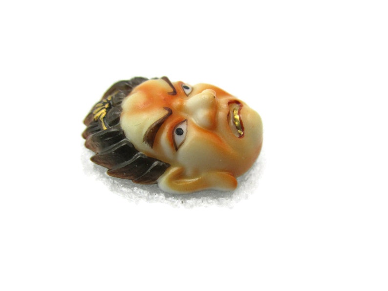 May include: A ceramic mask with a detailed face, painted in shades of orange and brown. The mask has black hair and gold accents around the eyes and mouth.