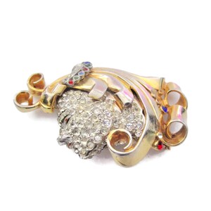 May include: A gold-tone brooch with a large, clear rhinestone cluster in the center. The brooch is decorated with swirls and scrolls, and has a few small red, blue, and green rhinestones.
