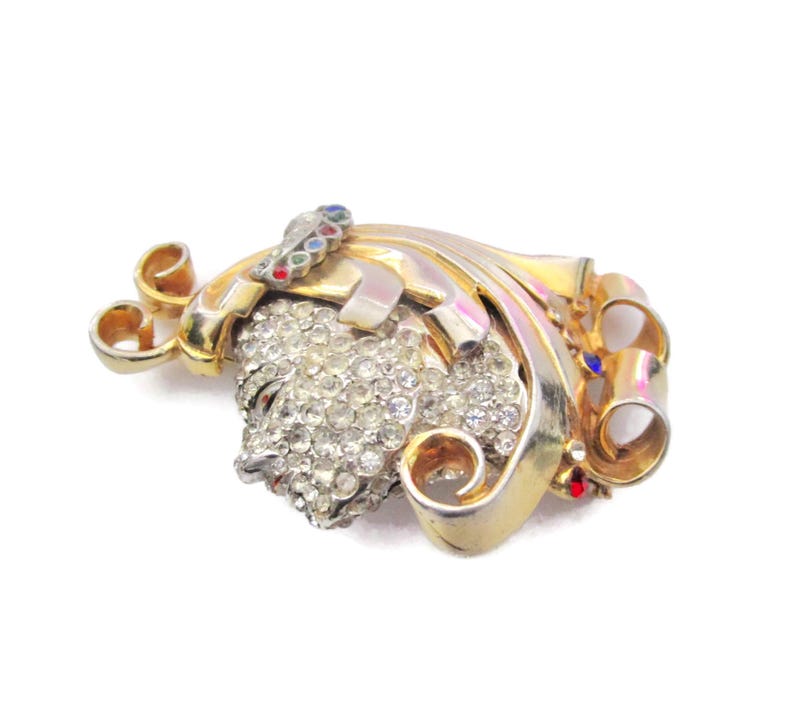 May include: A gold-tone brooch with a large, clear rhinestone cluster in the center. The brooch is decorated with swirls and scrolls, and has a few small red, blue, and green rhinestones.