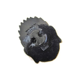 May include: Black metal pin with a detailed design. The back of the pin has the word "JAPAN" printed on it.