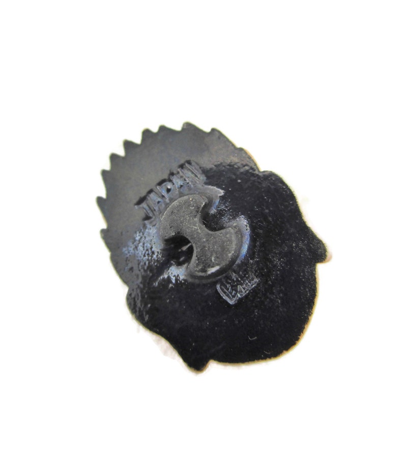May include: Black metal pin with a detailed design. The back of the pin has the word "JAPAN" printed on it.