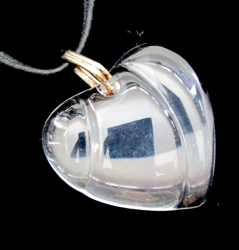 May include: A clear glass heart-shaped pendant with a gold-tone chain. The pendant is layered with a second heart shape, creating a dimensional effect.