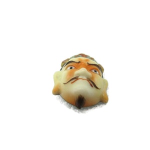 May include: A ceramic mask with a painted face, featuring a yellow and orange crown, black eyebrows, and a black mustache. The mask has a white background and a slightly frowning expression.