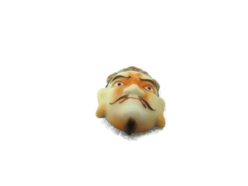 May include: A ceramic mask with a painted face, featuring a yellow and orange crown, black eyebrows, and a black mustache. The mask has a white background and a slightly frowning expression.