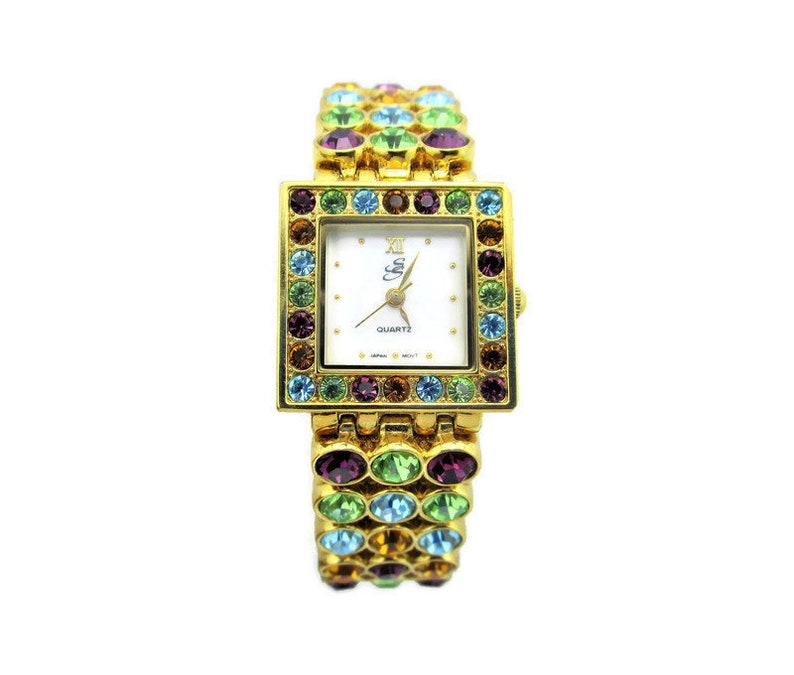 May include: A gold-tone watch with a white face and a crystal-encrusted band. The band is made up of small, colorful stones in shades of green, blue, purple, and orange. The watch face has black Roman numeral hour markers and the words "QUARTZ" and "Ella" printed on it.