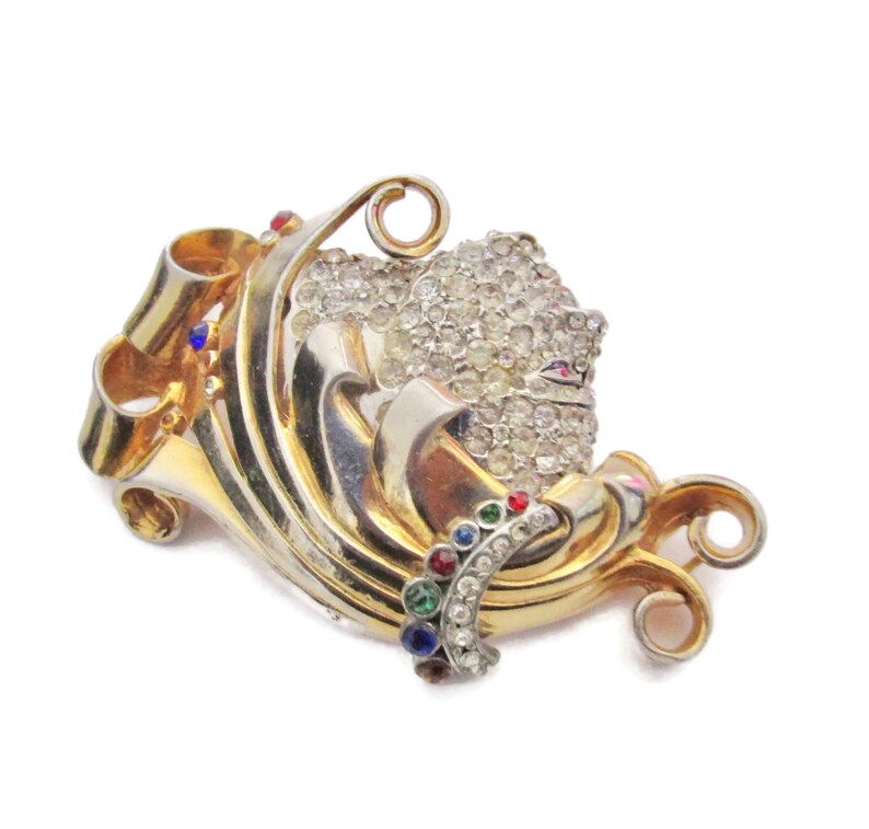 May include: A gold and silver tone brooch with a swirl design and a cluster of clear rhinestones. The brooch is accented with red, blue, and green gemstones.