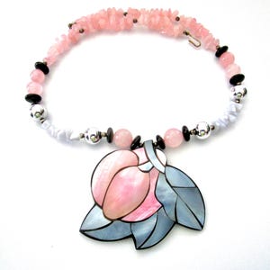 May include: A pink gemstone necklace with a floral pendant. The necklace features a strand of small pink stones, black beads, and silver-colored accents. The pendant is a stylized flower in shades of pink and gray.