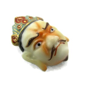 May include: A porcelain figurine of a man's face with a crown. The face is painted with details, including a mustache, eyebrows, and eyes. The crown is painted in red, blue, and gold.